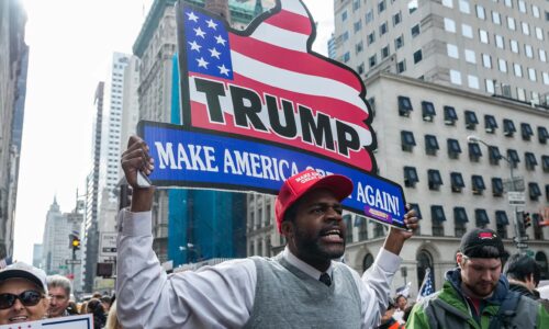 African American rally-goer wearing a Trump slogan hat boldly holds a patriotic-themed thumbs-up sign reading "Trump | Make America Great Again!"