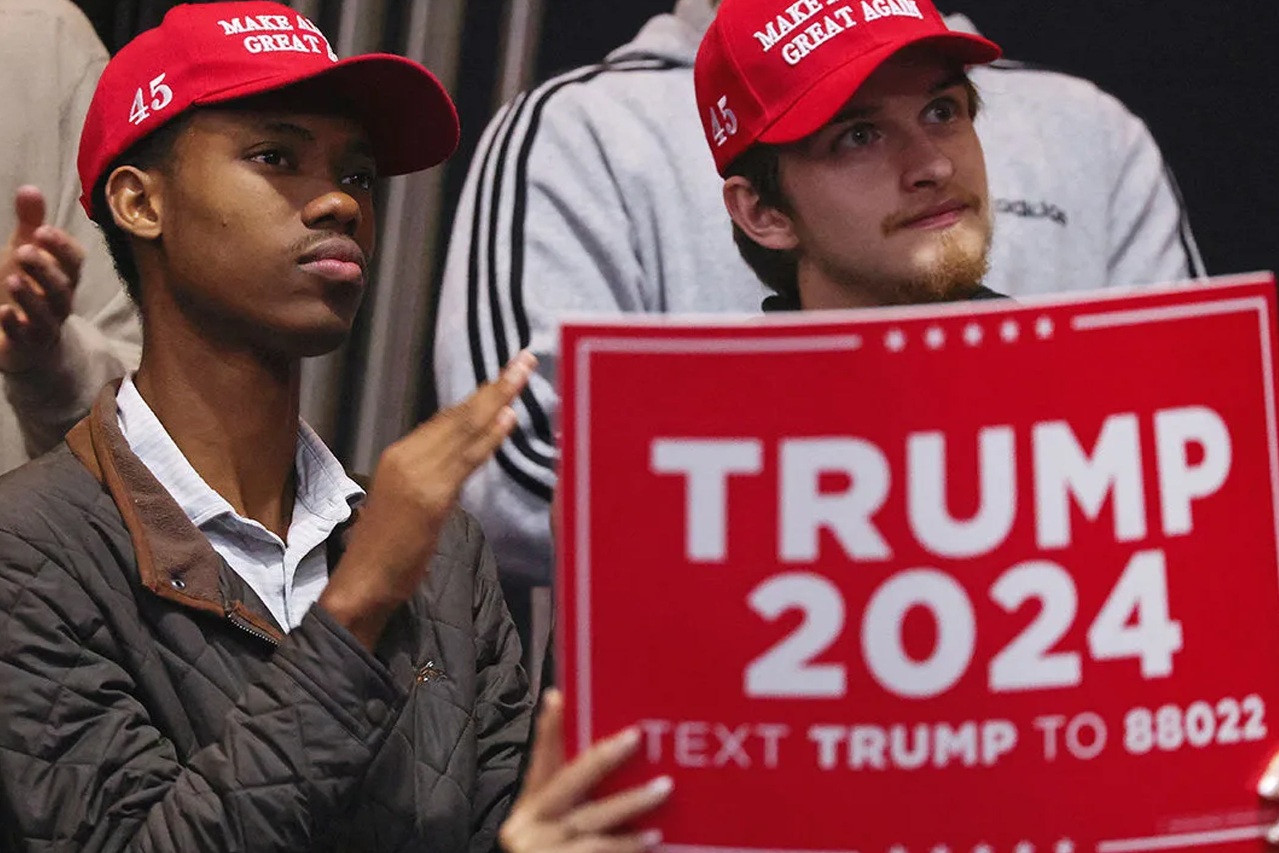 Young black and white voters, both wearing "MAGA" hats, sit side by side at a Trump 2024 rally intently watching the platform.